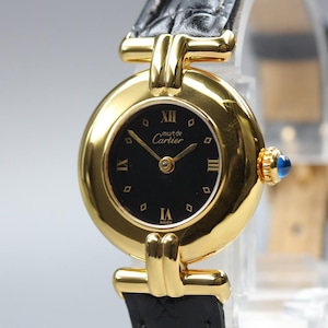 May include: A gold-tone wristwatch with a round case and a black dial featuring Roman numerals at the twelve, three, six, and nine o'clock positions. The dial displays the text 'must de Cartier' and 'Swiss'. It has a black textured leather strap.