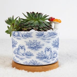 May include: A ceramic planter with a blue and white toile pattern featuring dogs and floral designs. The planter holds a succulent plant and decorative mushroom figurines. The planter sits on a wooden base.