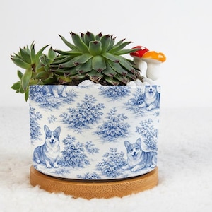 May include: A ceramic planter with a blue and white toile pattern featuring corgis and foliage. The planter holds a succulent plant, white pebbles, and decorative mushroom figurines. It sits on a round wooden base.
