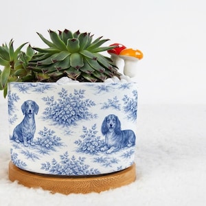 May include: A ceramic planter with a blue and white toile pattern featuring dachshunds. The planter holds a green succulent plant, white pebbles, and decorative mushroom figurines. It sits on a round wooden base.