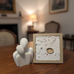 Könnte beinhalten: Eine weiße abstrakte Skulptur von zwei sich umarmenden Figuren, neben einer gerahmten 3D-Karte mit kreisförmigem Design. Die Karte ist weiß mit einem Holzrahmen. Die Objekte stehen auf einem Holztisch.