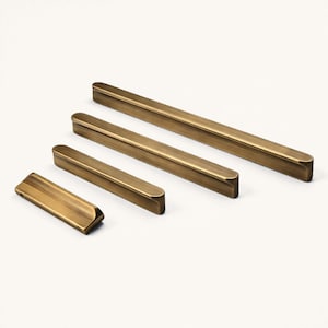 May include: Four brass-toned cabinet pulls of varying lengths are arranged on a white surface. The pulls are rectangular with rounded edges, and the finish has a subtle sheen. The sizes range from small to long, suggesting a set for different drawer or door sizes.