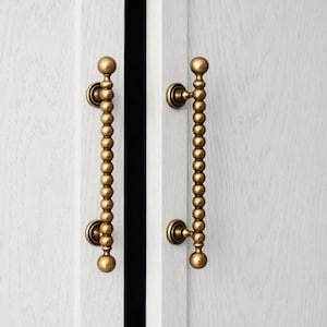 May include: Two gold-colored cabinet pulls with a beaded design. The pulls are mounted on a white cabinet door. The pulls have a vertical beaded design with a round knob at the top and bottom.