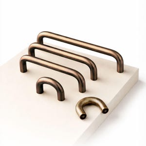 May include: A collection of bronze-colored cabinet pulls in various sizes. The pulls are curved, with a rounded shape, and are arranged on a white surface. The hardware is made of metal and has a vintage aesthetic.