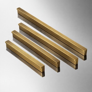 May include: Four brass cabinet pulls of different lengths are arranged diagonally. The pulls have a rectangular shape with a slightly raised edge on the ends. The brass finish has a warm, golden tone, and they are set against a reflective surface.