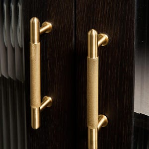 May include: Close-up of two gold cabinet handles. The handles are cylindrical with a textured, ridged design and are mounted on a dark brown wooden cabinet door. The handles have a modern, minimalist aesthetic.