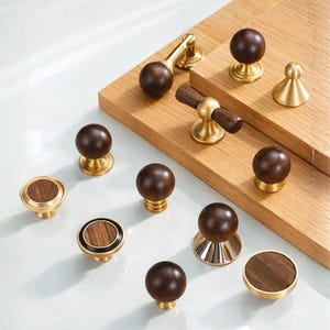 May include: A collection of various cabinet knobs and pulls. The hardware features a combination of dark wood and brass or gold-toned metal. Designs include round knobs, cylindrical pulls, and decorative accents.