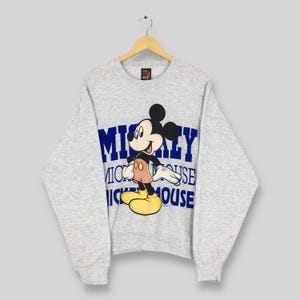 K&ouml;nnte beinhalten: Hellgraues Sweatshirt mit Mickey Mouse-Grafik. Die Zeichentrickfigur ist farbig, mit einem blauen "Mickey Mouse"-Textdesign. Das Sweatshirt h&auml;ngt an einem Holzb&uuml;gel.