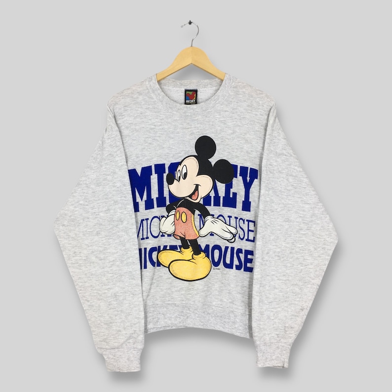 K&ouml;nnte beinhalten: Hellgraues Sweatshirt mit Mickey Mouse-Grafik. Die Zeichentrickfigur ist farbig, mit einem blauen "Mickey Mouse"-Textdesign. Das Sweatshirt h&auml;ngt an einem Holzb&uuml;gel.