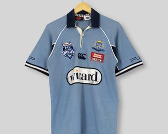 Puede incluir: Polo de color azul claro con cuello azul marino y detalles blancos. La camiseta presenta varios parches, incluyendo "Wizard", "NSW ORIGIN" y "Harvey Norman Fujifilm". Tiene mangas cortas con puños a rayas azul marino y blanco.