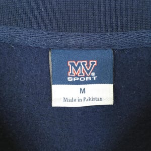 Op de afbeelding: Close-up van een marineblauw kledingstuk met een label. Het label bevat de tekst "MV SPORT" in rood en wit, met de letter "M" eronder. Op het label staat ook "Made in Pakistan".