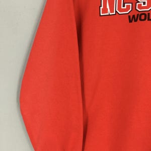 May include: Bright orange sweatshirt with white and black text that reads "NC STATE WOLVES". The sweatshirt is a solid color with a crew neck and long sleeves. The text is in a bold, blocky font.