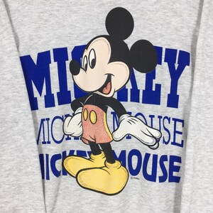 K&ouml;nnte beinhalten: Ein graues Sweatshirt mit einer Mickey Mouse-Grafik. Das Design enth&auml;lt die Worte "Mickey Mouse" in blauen Blockbuchstaben. Mickey wird in einer stehenden Pose dargestellt und tr&auml;gt rote Shorts, gelbe Schuhe und wei&szlig;e Handschuhe.