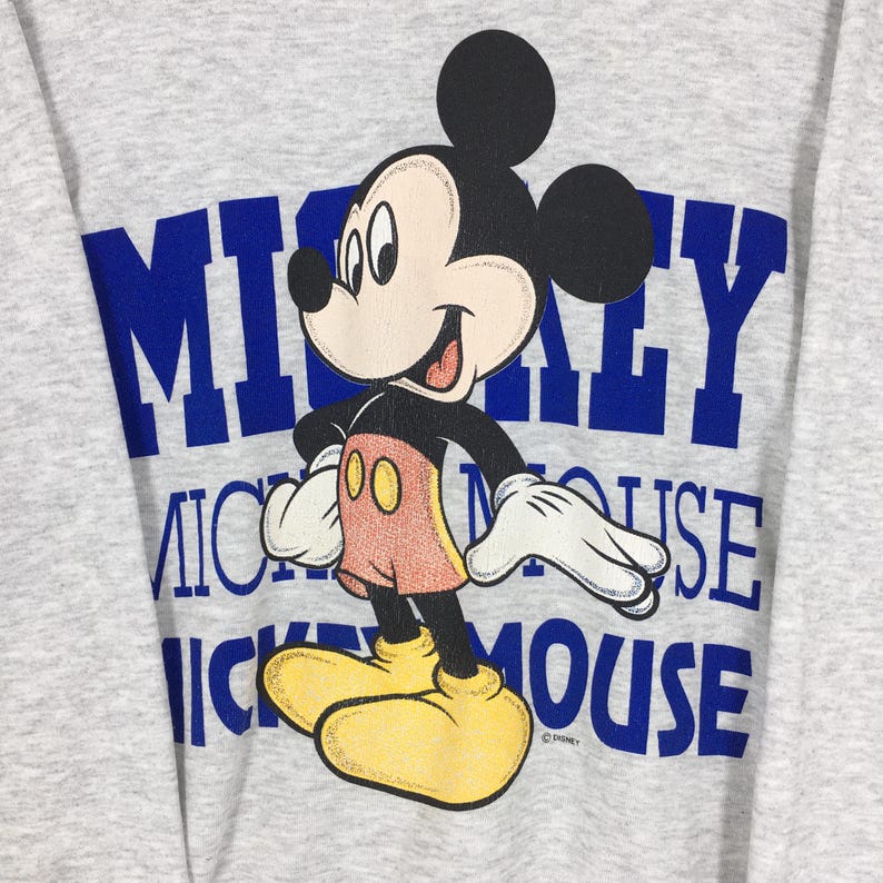 K&ouml;nnte beinhalten: Ein graues Sweatshirt mit einer Mickey Mouse-Grafik. Das Design enth&auml;lt die Worte "Mickey Mouse" in blauen Blockbuchstaben. Mickey wird in einer stehenden Pose dargestellt und tr&auml;gt rote Shorts, gelbe Schuhe und wei&szlig;e Handschuhe.