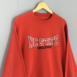 May include: A red NC State Wolfpack sweatshirt. The sweatshirt has the words "NC STATE" in white block letters with a black outline, and "WOLFPACK" in smaller white letters below. The sweatshirt is hanging on a wooden hanger.