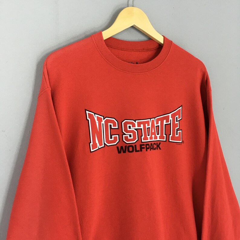 May include: A red NC State Wolfpack sweatshirt. The sweatshirt has the words "NC STATE" in white block letters with a black outline, and "WOLFPACK" in smaller white letters below. The sweatshirt is hanging on a wooden hanger.