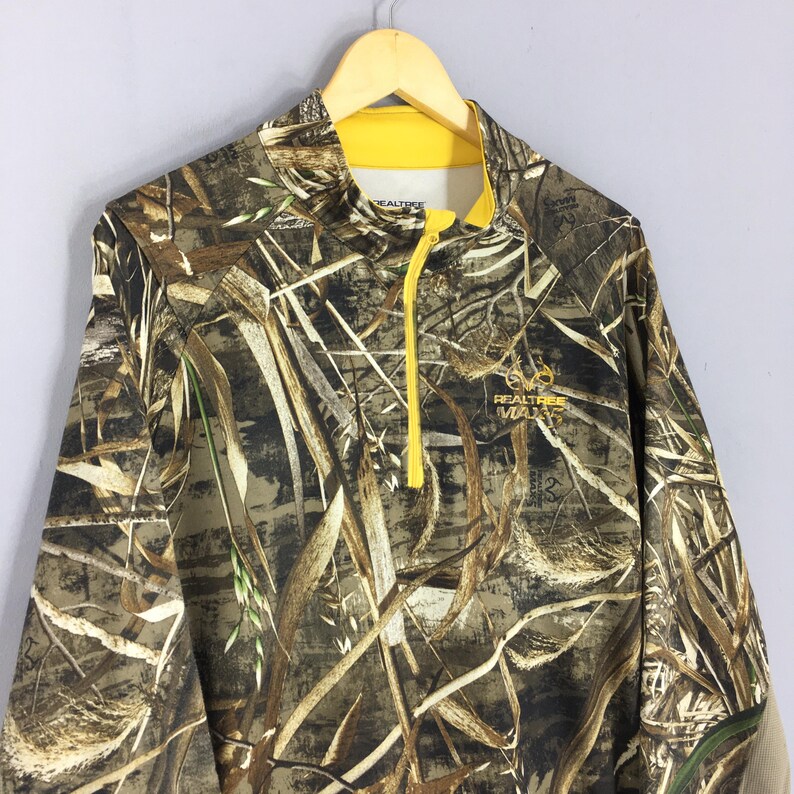 Puede incluir: Una camisa de camuflaje de manga larga con cremallera y cuello amarillos. La camisa presenta un logotipo de Realtree Max-3 y un patr&oacute;n de colores marr&oacute;n, verde y beige, que se asemeja a un entorno natural al aire libre. La camisa est&aacute; colgada en una percha de madera.