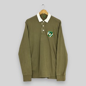 May include: Olive green long-sleeve polo shirt with a white collar and button placket. Features a Benetton logo patch on the chest. The shirt is hanging on a wooden hanger against a light gray background.