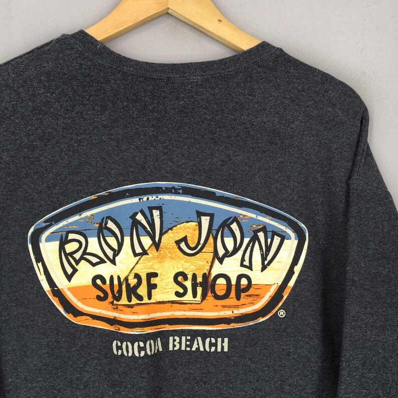 May include: Charcoal grey sweatshirt with a distressed oval logo. The logo features the text "RON JON SURF SHOP" in white and yellow, set against a blue, orange, and brown background. The words "COCOA BEACH" are printed below the logo.