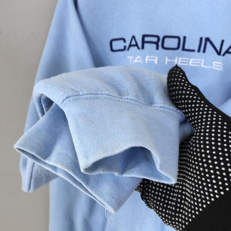 Puede incluir: Sudadera azul claro con el texto "CAROLINA TAR HEELS" en azul marino. Se ve el pu&ntilde;o de la manga, sostenido por una mano con un guante negro con lunares blancos. La sudadera es de un material suave.