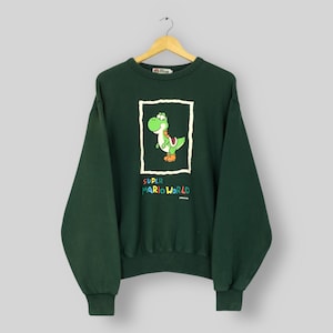 May include: Dark green sweatshirt featuring a graphic of Yoshi from Super Mario World. The image of Yoshi is in a white rectangle, with the words "SUPER MARIO WORLD" below. The sweatshirt has a crew neck and long sleeves.