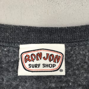 May include: A close-up shot of a dark grey sweatshirt with a white label. The label displays the words "RON JON SURF SHOP" in red and orange lettering, outlined in black, on a white background. The sweatshirt has a textured appearance.