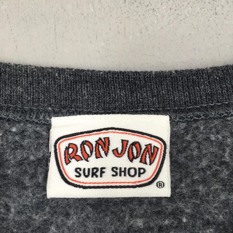 May include: A close-up shot of a dark grey sweatshirt with a white label. The label displays the words "RON JON SURF SHOP" in red and orange lettering, outlined in black, on a white background. The sweatshirt has a textured appearance.