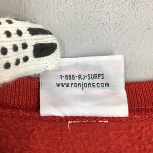 May include: A close-up shot of red fabric with a white label. The label displays the text "1-888-RJ-SURFS" and "www.ronjons.com" in black. A section of a white and black patterned item is visible in the top left corner.