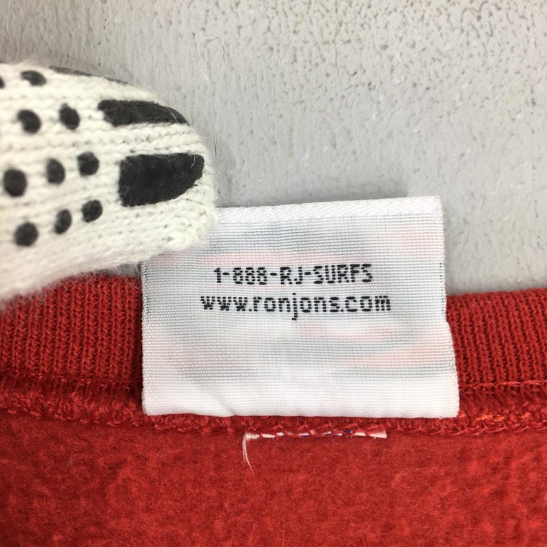 May include: A close-up shot of red fabric with a white label. The label displays the text "1-888-RJ-SURFS" and "www.ronjons.com" in black. A section of a white and black patterned item is visible in the top left corner.