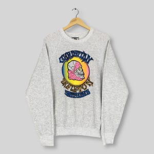 May include: A light gray sweatshirt with a crew neck and long sleeves. The front features a graphic of a skull wearing a pink bandana, surrounded by text that reads "CHRISTIAN SKELETON SKATEBOARDS" in blue and gold.