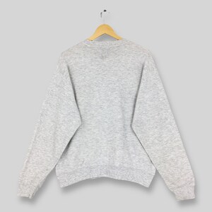 K&ouml;nnte beinhalten: Ein melierter grauer Pullover h&auml;ngt an einem Holzkleiderb&uuml;gel. Das lang&auml;rmlige Oberteil hat einen Rundhalsausschnitt und gerippte B&uuml;ndchen. Die R&uuml;ckseite des Sweatshirts ist sichtbar und zeigt den einfarbigen Stoff. Das Kleidungsst&uuml;ck scheint aus einem weichen, bequemen Material zu sein.