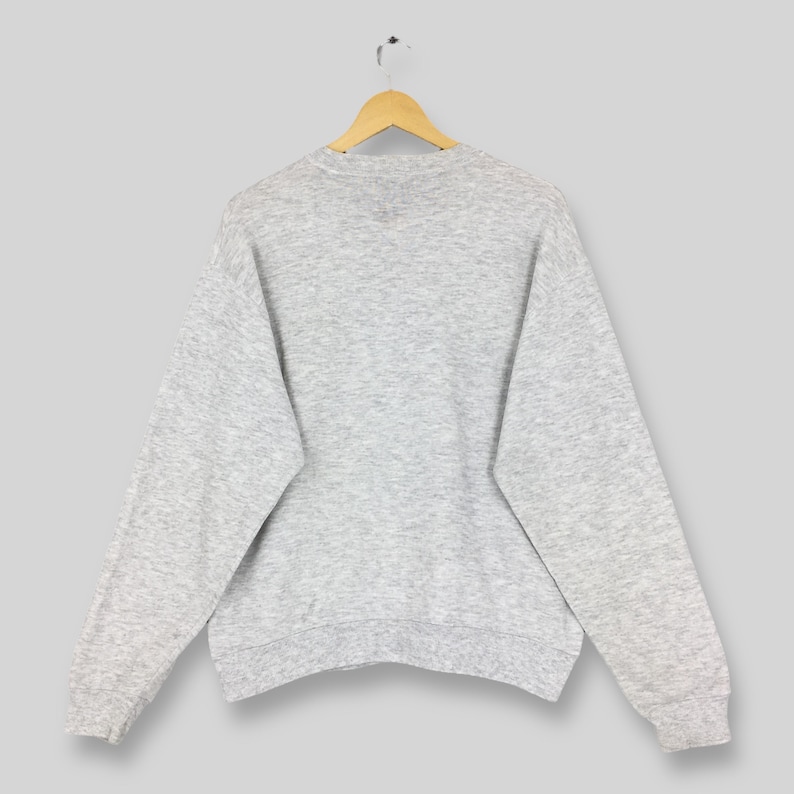 K&ouml;nnte beinhalten: Ein melierter grauer Pullover h&auml;ngt an einem Holzkleiderb&uuml;gel. Das lang&auml;rmlige Oberteil hat einen Rundhalsausschnitt und gerippte B&uuml;ndchen. Die R&uuml;ckseite des Sweatshirts ist sichtbar und zeigt den einfarbigen Stoff. Das Kleidungsst&uuml;ck scheint aus einem weichen, bequemen Material zu sein.