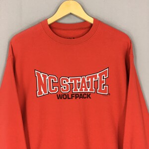 May include: A red sweatshirt with the text "NC STATE WOLFPACK" in white and black lettering. The sweatshirt is hanging on a wooden hanger.