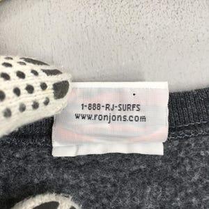 May include: A close-up of a grey garment label with the text "1-888-RJ-SURFS" and "www.ronjons.com" printed in black. The label is white with a red stripe at the bottom. The garment is a dark grey colour.