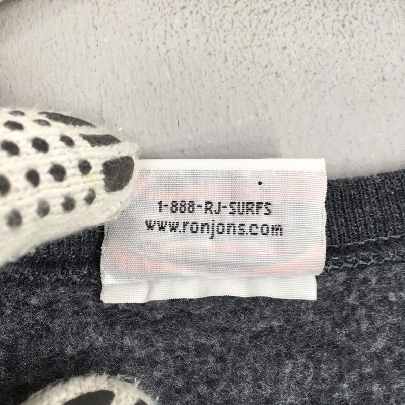 May include: A close-up of a grey garment label with the text "1-888-RJ-SURFS" and "www.ronjons.com" printed in black. The label is white with a red stripe at the bottom. The garment is a dark grey colour.