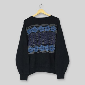 May include: Black crew neck sweatshirt with a graphic design. The design features the text "MVU SPORTS" in a blue font, framed by blue floral patterns. The sweatshirt is hanging on a wooden hanger.