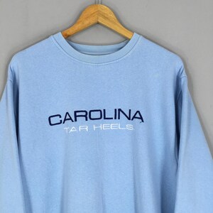 Puede incluir: Sudadera azul claro con la palabra "CAROLINA" bordada en azul marino en el pecho, con "TAR HEELS" debajo. La sudadera est&aacute; colgada en una percha de madera.