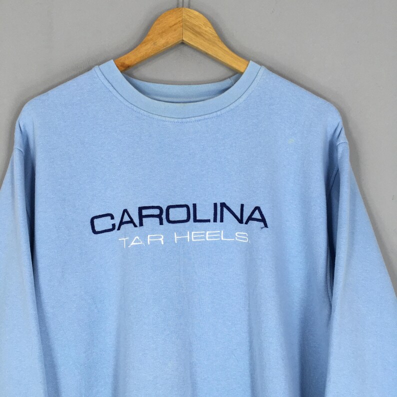 Puede incluir: Sudadera azul claro con la palabra "CAROLINA" bordada en azul marino en el pecho, con "TAR HEELS" debajo. La sudadera est&aacute; colgada en una percha de madera.