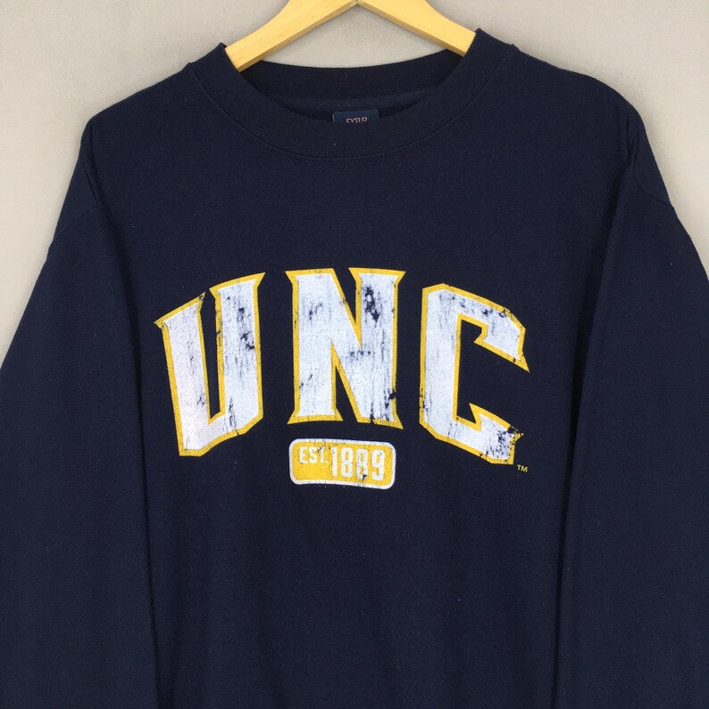 Op de afbeelding: Marineblauwe sweatshirt met de letters "UNC" in geel en wit, met "EST 1889" eronder. De sweatshirt hangt aan een houten hanger.