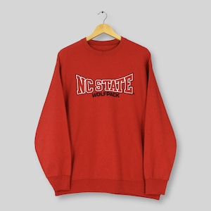 May include: A red crewneck sweatshirt with the text "NC STATE WOLFPACK" in white and black lettering. The sweatshirt is hanging on a wooden hanger against a light background. The sweatshirt has long sleeves and a ribbed neckline.