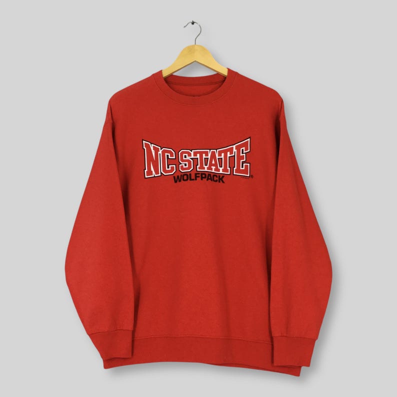 May include: A red crewneck sweatshirt with the text "NC STATE WOLFPACK" in white and black lettering. The sweatshirt is hanging on a wooden hanger against a light background. The sweatshirt has long sleeves and a ribbed neckline.