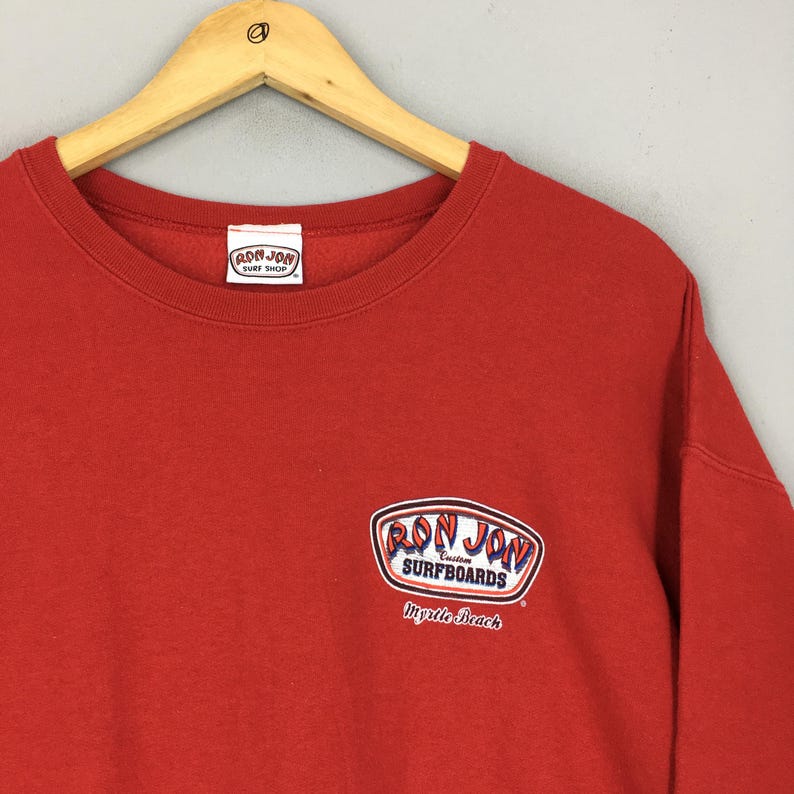 May include: A red crew neck sweatshirt featuring the Ron Jon Surf Shop logo on the upper left chest and a larger Ron Jon Surfboards logo on the lower right. The sweatshirt is hanging on a wooden hanger.