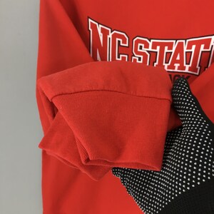 May include: A close-up of a red sweatshirt sleeve with the text "NC STATE" in white block letters outlined in black. The sleeve is folded over, revealing the cuff. A black and white dotted glove is in the image.