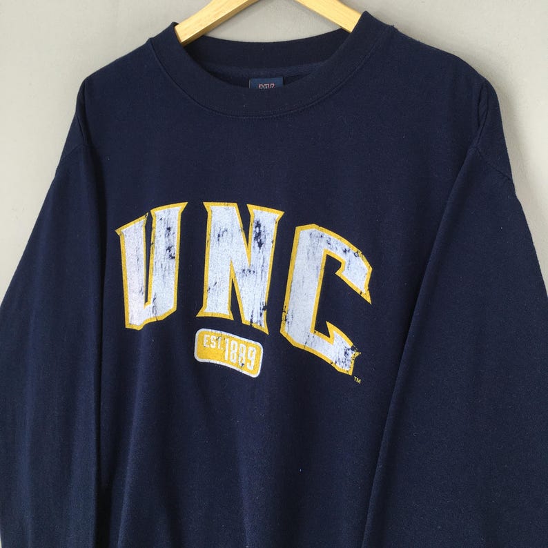 Op de afbeelding: Marineblauw sweatshirt met de letters "UNC" in wit en geel, met de tekst "EST 1889" eronder. Het sweatshirt hangt aan een houten kleerhanger.