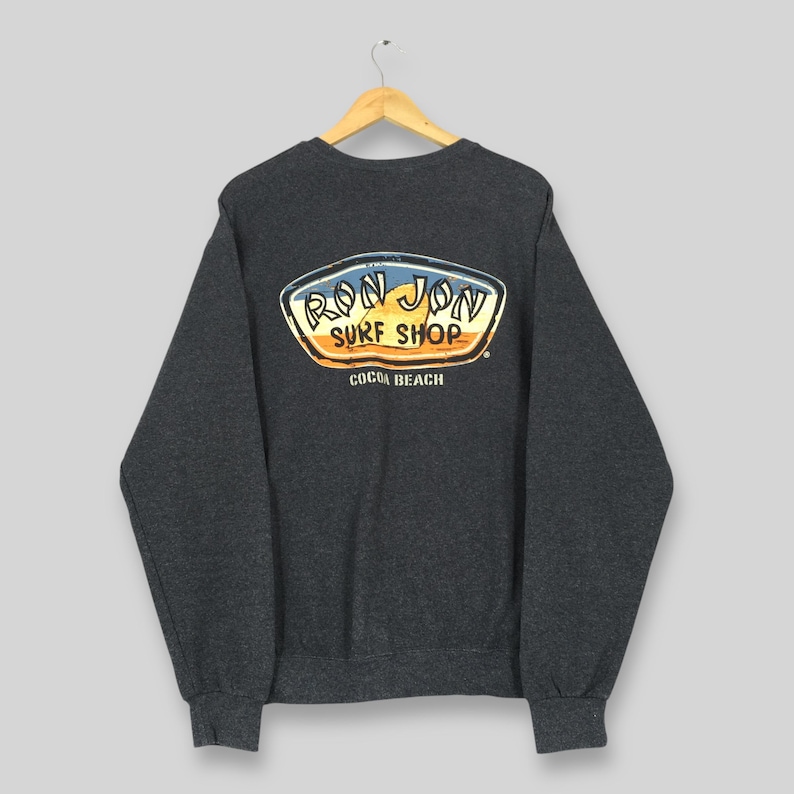 May include: A dark grey sweatshirt featuring the Ron Jon Surf Shop logo on the back. The logo is a colourful design with the words "Ron Jon Surf Shop" and "Cocoa Beach". The sweatshirt has a crew neck and long sleeves.