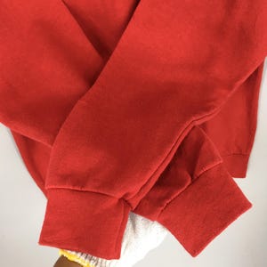 May include: Close-up of a red sweatshirt. The sleeves are folded over each other, with the cuffs visible. The fabric appears soft and slightly textured. The cuffs are ribbed and a slightly darker shade of red.