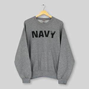 Könnte beinhalten: Ein graues Sweatshirt mit dem Wort "NAVY" in großen, schwarzen Buchstaben auf der Brust. Das Sweatshirt hängt an einem Holzbügel vor einem hellgrauen Hintergrund. Es hat lange Ärmel und einen Rundhalsausschnitt.
