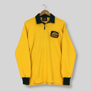 Könnte beinhalten: Ein langärmeliges gelbes Rugby-Shirt mit dunkelgrünem Kragen und Manschetten. Das Shirt hat ein kleines Emblem mit dem australischen Wappen auf der Brust. Das Shirt hängt an einem Holzkleiderbügel.