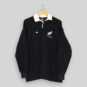 May include: Black long-sleeve rugby shirt with a white collar. The shirt features the Canterbury logo and a white fern with "NEW ZEALAND" printed below it. The shirt is hanging on a wooden hanger.