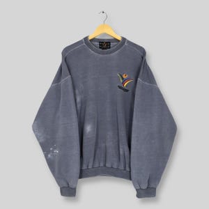 May include: A vintage, faded blue sweatshirt with a crew neck and ribbed cuffs and hem. The sweatshirt features a colourful embroidered logo on the chest, with the text "4th World Masters Games". The garment is hanging on a wooden hanger.